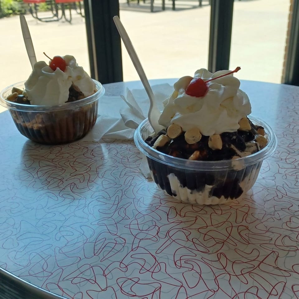 Two chocolate ice cream sundaes topped with whipped cream, cherries, and nuts sit on a patterned café table.