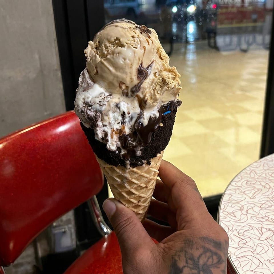 Hand holding a waffle cone topped with chocolate cake, vanilla ice cream, chocolate drizzle, and cookie crumbles inside a dessert shop.