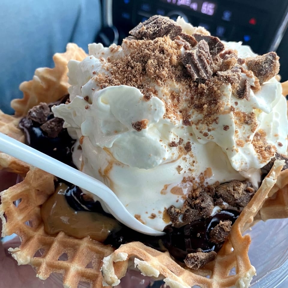 Waffle bowl sundae topped with whipped cream, chocolate pieces, cookie crumbs, caramel, and chocolate syrup.