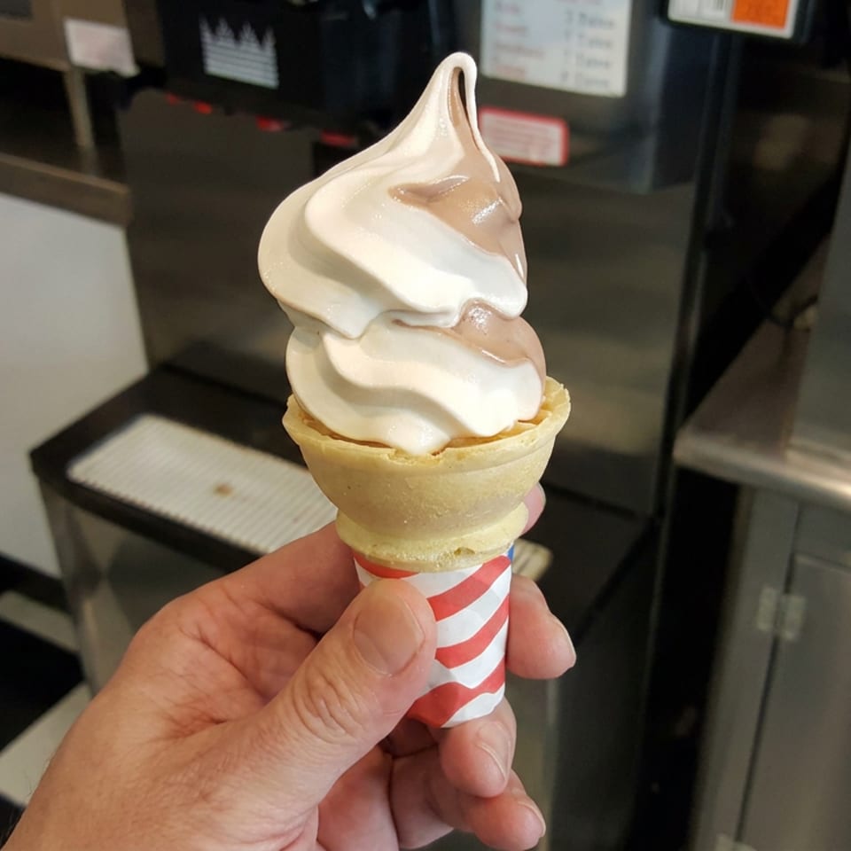 Hand holding a red-and-white striped cone topped with two swirls of vanilla soft-serve ice cream inside a restaurant.